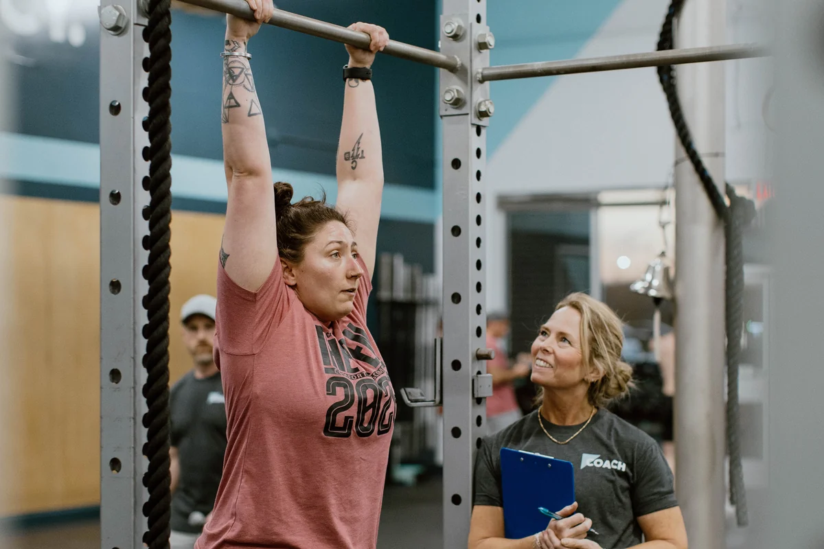 A CrossFit Spring Hill member hanging from a pull-up bar while a coach stands beside her watching her setup and taking notes