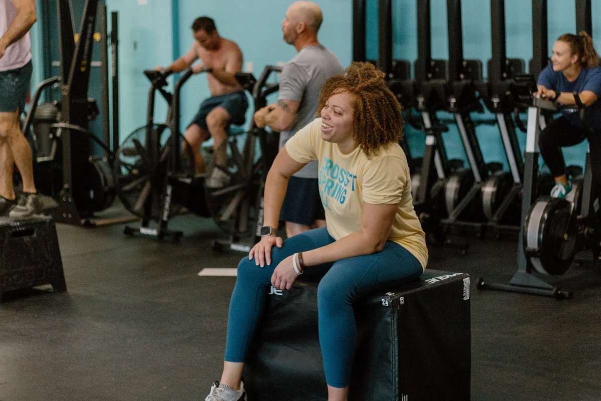 A CrossFit Spring Hill member sitting on a box laughing during a break while class continues behind her