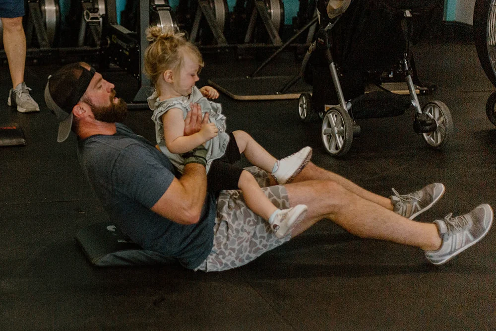 A dad doing sit-ups at CrossFit Spring Hill with his toddler daughter held on his chest, stroller parked beside him
